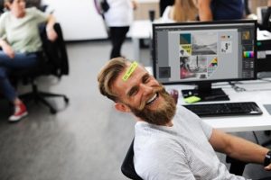 Young man with a beard smiling at his work desk with "be happy" written on his forehead