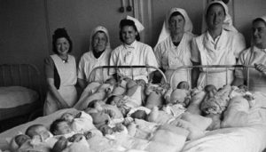 Nurses standing around many babies on a hospital bed in WW2